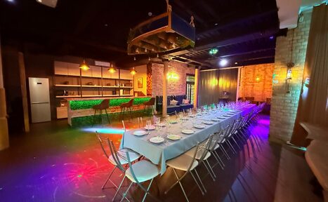Арендовать Лофт Фантом. Пространство для мероприятий с длинным столом, накрытым скатертью и сервированным посудой, рядом складные стулья. На заднем плане барная стойка с высокими стульями. Кирпичные стены, потолок с открытыми балками. Разноцветное освещение.