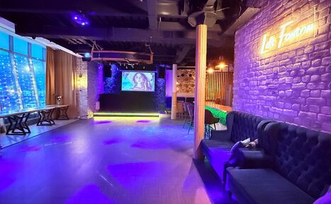 Арендовать Лофт Фантом. Пространство в стиле лофт с кирпичными стенами, неоновыми светильниками и деревянными элементами. На стене проектор, рядом с диваном стулья. Быстрые столы вдоль окна, занавешенные шторами. Потолочные светильники создают разноцветное освещение.