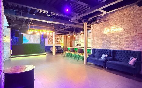 Арендовать Лофт Фантом. Просторное помещение с открытой планировкой, кирпичные стены, барная стойка с высокими стульями, синий диван, неоновая вывеска, большой экран для проекций. Потолок темный с открытыми балками, комбинированное освещение. Пол плиточный.