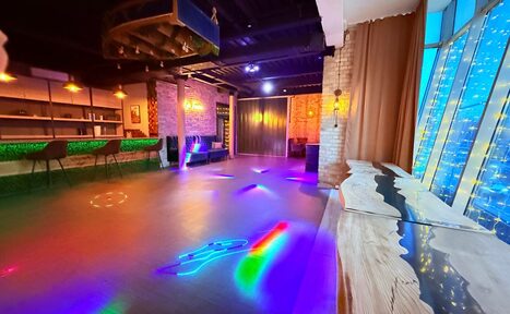 Арендовать Лофт Фантом. Просторное помещение с барной стойкой, высокими табуретами и открытыми полками. На полу разноцветные световые эффекты. Окна украшены светодиодными гирляндами. Стены из кирпича, потолок темный, длинный деревянный стол у окна.