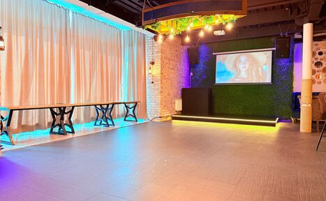 Арендовать Лофт Фантом. Просторное помещение с занавесками, кирпичными стенами и светлым плиточным полом. Включает три деревянных стола с металлическими ножками, проектор на стене с зеленым покрытием, яркое разноцветное освещение и декоративные элементы на потолке.