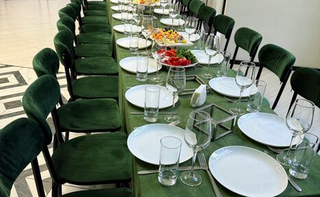 Арендовать Arcobaleno Loft(Аркобалено Лофт). Длинный стол, накрытый скатертью зелёного цвета, окружён стульями с зелёной обивкой. На столе расставлены белые тарелки, бокалы и стаканы, приборы. Разложены салаты и овощи. Пол выложен плиткой с черно-белым рисунком. Освещение естественное.