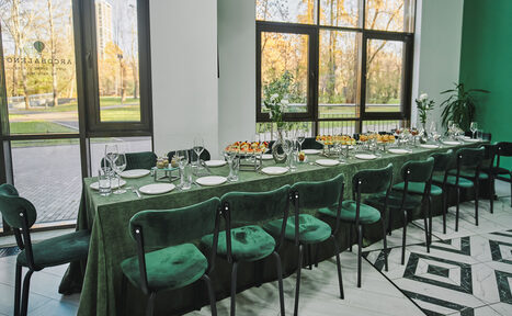 Арендовать Arcobaleno Loft(Аркобалено Лофт). Просторное помещение для мероприятий с длинным столом, покрытым зелёной скатертью, и зелёными стульями. На столе посуда и закуски. Широкие окна с видом на деревья. Черно-белый узор на полу. Светлые стены.