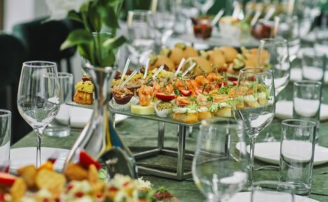 Арендовать Arcobaleno Loft(Аркобалено Лофт). Длинный обеденный стол, покрытый зелёной скатертью, с расставленными белыми тарелками и стеклянной посудой. В центре стола на подставке расположено разнообразное канапе. На заднем плане видна зелень в вазе.