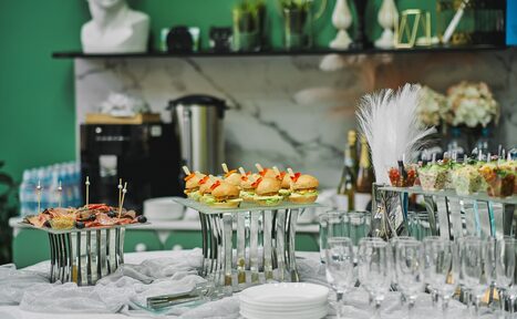 Арендовать Arcobaleno Loft(Аркобалено Лофт). Стол с закусками на подставках, ряды бокалов и белые тарелки. Задний план: кофемашина, бутылки, мраморная стена, декор, зелёные стены, вазы и статуэтка.