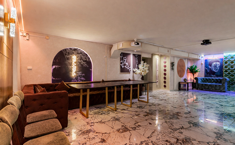 Арендовать Стильный лофт Джаванди. Просторное помещение со светлым мраморным полом, длинным темным столом и мягкими креслами. Стены украшают картины и декоративные панели. Разное освещение: потолочные светильники и неоновая подсветка. Украшено растениями и цветами в вазах.