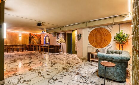 Арендовать Стильный лофт Джаванди. Просторное помещение с мраморным полом. В центре — изогнутый диван и круглый столик. Стены украшены настенной графикой с кругами. Барная стойка и высокие стулья у стены. Потолочное освещение и проектор. Декоративные растения в вазах.