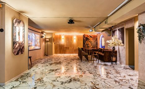 Арендовать Стильный лофт Джаванди. Мраморный пол с разноцветными прожилками, нейтральные стены, деревянная панель на дальней стене, несколько стульев и стойка, проектор на потолке, настенные картины и декор, мягкое освещение, крупная ваза с белыми цветами.