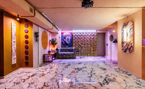 Арендовать Стильный лофт Джаванди. Пространство с мраморным полом, кожаным диваном у стены с декоративными тарелками и неоновой подсветкой. Потолок светлый, светильники на стенах и потолке. Колонны украшены панелями и декоративными элементами. Стены светлых тонов.