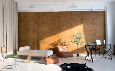 Арендовать Зал Серебро. Просторная и светлая фотостудия. Просторное помещение с белыми стенами и полом, акцентированное деревянной панелью. Мебель: белый диван, коричневые пуфы, черный стол, зеленый стул. Освещение естественное. Декор: растение в горшке, ковёр с животным принтом. Справа ноутбук на столе.