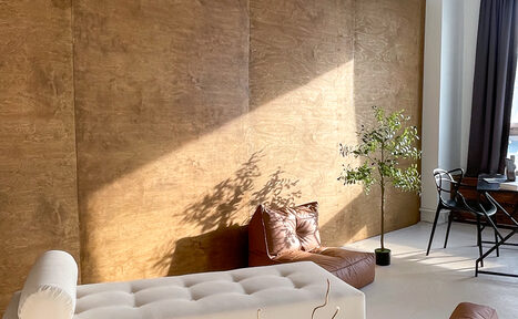 Арендовать Зал Серебро. Просторная и светлая фотостудия. Пространство с деревянной стеной, белым диваном и коричневой кожаной подушкой. У окна черный металлический стол с креслом, рядом зеленое растение в горшке. Естественное освещение, светло-серая штора, пол светлого оттенка.