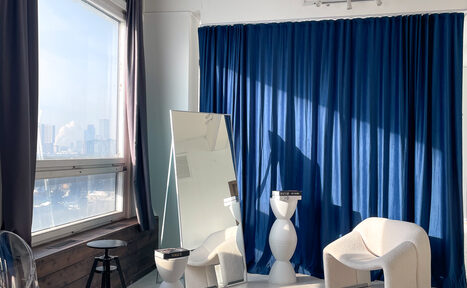 Арендовать Зал Серебро. Просторная и светлая фотостудия. Пространство со светлым креслом, высоким прямоугольным зеркалом, круглым белым столиком с книгами, тёмно-синей драпировкой на стене. Большое окно с серыми шторами, виден городской пейзаж. Слева деревянный стул, стены и пол светлые. Естественное освещение.