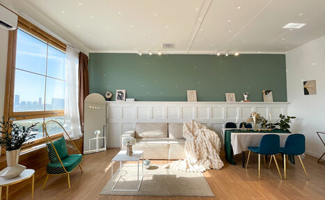 Арендовать Зал Квартира. Уютное пространство в квартирном стиле. Светлая комната с деревянным полом и зелёной акцентной стеной. Белый диван, зеркало в полный рост, небольшой столик, обеденный стол с синими стульями. Большое окно, шторы. Размещение предметов декора на полке. Подвесное освещение.