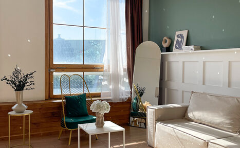 Арендовать Зал Квартира. Уютное пространство в квартирном стиле. Гостиная с большим окном, бело-зелеными стенами и деревянным полом. Мебель: бежевый диван, белый стол, золотистое кресло с зеленой подушкой. Декор: зеркало, вазы с цветами, книги. Естественное освещение, белые шторы, деревянные элементы.