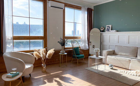 Арендовать Зал Квартира. Уютное пространство в квартирном стиле. Гостиная с деревянным полом, двумя большими окнами и бежевыми шторами. Светлый диван, два кресла, круглые столики. Стена зеленого цвета с деревянными панелями, есть декор в виде сухоцветов. Большое зеркало и кондиционер на стене. Естественное освещение.