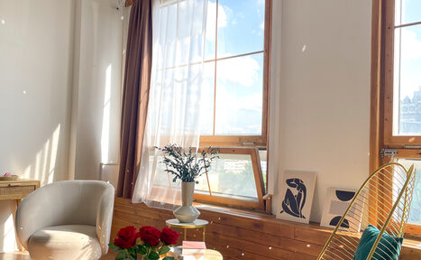 Арендовать Зал Квартира. Уютное пространство в квартирном стиле. Светлое помещение с большими окнами и длинными шторами, белое кресло, деревянный стол с букетом красных роз, декоративные картины на подоконнике, растение в вазе, деревянные элементы, светлый текстиль.