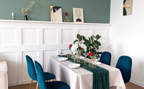 Арендовать Зал Квартира. Уютное пространство в квартирном стиле. Столовая зона с прямоугольным столом, накрытым белой скатертью и зелёной дорожкой. Синие стулья, белые панели на стене, зелёная стена сверху, картины и декор на полке. Светлое помещение с деревянным полом и декоративными растениями.