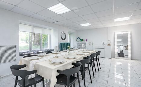 Арендовать Handy Class (Хэнди Класс). Современный зал. Просторная светлая комната с большим обедом столом, за которым расположены черные стулья. На столе бежеватая скатерть, посуда и декоративные свечи. Потолок с квадратными светильниками, пол выложен белой плиткой. Возле окна мягкие подушки.