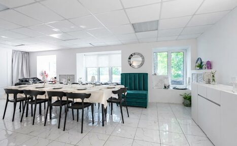 Арендовать Handy Class (Хэнди Класс). Современный зал. Просторное светлое помещение с белым плиточным полом и потолочной плиткой. Большой стол с черными стульями в центре, накрытый светлой скатертью. На стене круглое зеркало. Из окон виден зелёный пейзаж. В углу зелёный диван и белая тумба.