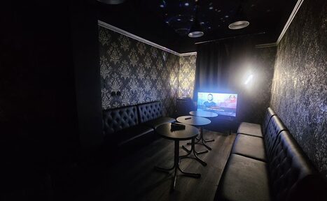 Арендовать Типичный Питер. ВИП лофт -зал. Тёмная комната с кожаными диванами вдоль стен, телевизором на тумбе в углу. Стены оклеены обоями с узором. Небольшие круглые столы перед диванами. Потолок тёмный с точечным освещением. Справа чёрная штора. Освещение приглушенное.
