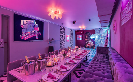 Арендовать Неоновый лофт Гламур. Караоке-зал с длинным обеденным столом, сервированным розовыми салфетками и приборами. Слева стена с панно, справа — длинный мягкий диван. Розовая неоновая надпись на стене, декоративная лампа и изображения на стенах. Свет фиолетовый и тёплый.
