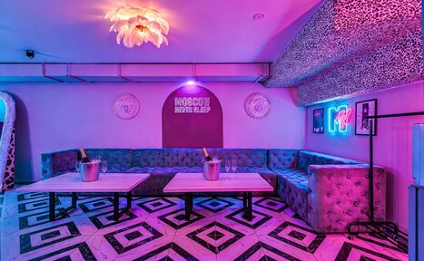 Арендовать Неоновый лофт Гламур. Клубное пространство с фиолетовым освещением, размещены мягкие угловые диваны серого цвета, два низких прямоугольных стола. Пол выложен плиткой с геометрическим рисунком. На стене неоновая вывеска, вокруг декор с леопардовым узором.