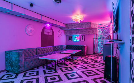Арендовать Неоновый лофт Гламур. Пространство с мягкими диванами вдоль стен, два стола. Пол с геометрическим узором. Неоновое освещение, фиолетовые и розовые тона. На стенах декоративные элементы, телевизор, неоновая вывеска. Подвесной светильник, звукозаписывающее оборудование.