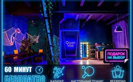 Арендовать Тематический лофт лагуна. Пространство с высоким потолком и темным интерьером, украшенное сетками и растениями. На стене неоновая надпись "Ocean Vibe". Мебель включает диван и столы. Неоновая вывеска "Подарок на выбор", освещение разноцветное с акцентом на синий. Текст: "60 минут бесплатно", "Караоке", "Настольный теннис", "Beerpong".