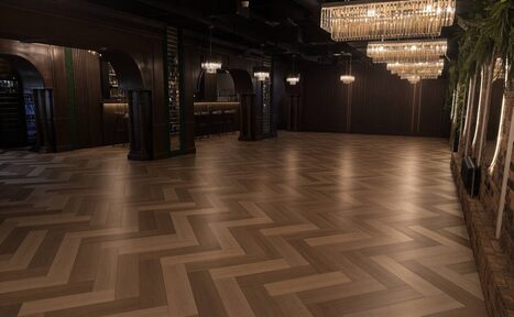 Арендовать Soul Event (Соул Ивент). Малый зал. Просторный зал с паркетным полом елочкой и темными деревянными стенами. Слева барная стойка с арочными проходами. Потолок темный с открытыми балками. Освещение с помощью больших люстр и светильников. На стенах живые растения.