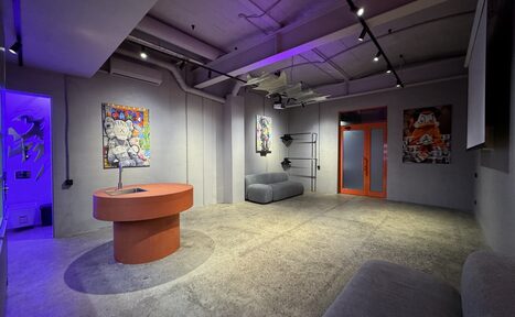 Арендовать Дизайнерский лофт-зал. Лофт-пространство с серыми бетонными стенами и полом, центральным круглым оранжевым столом, мягкими серыми диванами вдоль стен. Потолочное трековое освещение, несколько ярких картин, стеклянная дверь с оранжевой рамой.