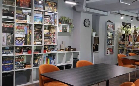 Арендовать Антикафе площадью 60 м² до 30 гостей. Современное помещение с полками, заполненными настольными играми. Столы с темными столешницами и оранжевыми стульями. Серые стены с часами, осветительные трек-светильники. Зеленые растения в горшках на полках. Индустриальный стиль с открытыми вентиляционными трубами.