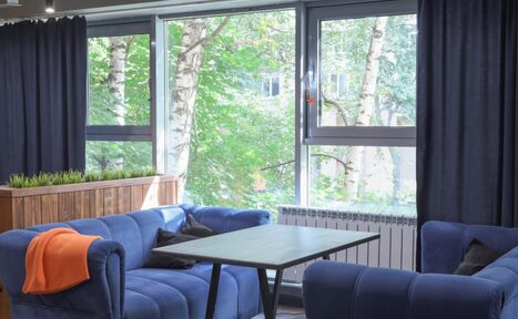 Арендовать Антикафе площадью 60 м² до 30 гостей. Современное помещение с большими окнами, темными шторами, синими диванами и черным столом. За окнами видны деревья. На диване оранжевый плед. Мебель расположена вокруг стола. На подоконнике деревянная панель с растениями.