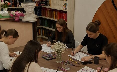 Арендовать Творческая лофт-студия под встречи и мастер-классы. Женщины сидят за столом в комнате с деревянными элементами и книжными полками. Стол украшен вазой с цветами, на нем лежат альбомы и карандаши. На заднем плане бюст и разные книги. Освещение мягкое, стены светлые.