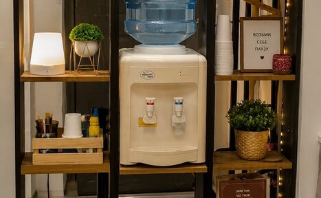 Арендовать Творческая лофт-студия под встречи и мастер-классы. Кулер с водой на полке, окружённой деревянными стеллажами. На полках лампа, стаканчики, пластиковые чашки, зелёные растения, контейнер с кофе-брейк принадлежностями. Рядом текст: «Возьми себе паузу». Мягкое освещение, тёмная рама стеллажа.