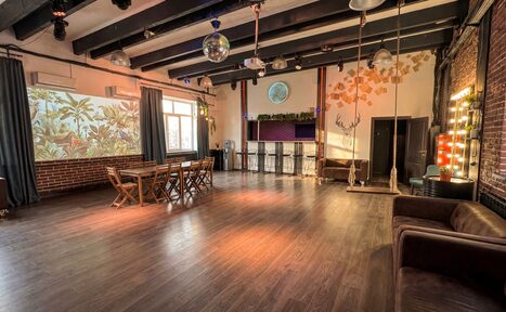 Арендовать Лофт с верандой, мангалом и сценическим оборудованием. Большое просторное помещение с деревянным полом. Стены из кирпича и штукатурки, частично декорированы рисунками листьев. Мебель: деревянный стол и стулья, барная стойка с высокими стульями, два дивана. Светильники подвесные, зеркальная шаровая лампа.