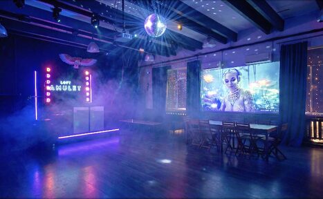 Арендовать Лофт с верандой, мангалом и сценическим оборудованием. Пространство с деревянным полом, столами и стульями. Неоновая вывеска с надписью "LOFT", зеркальный шар на потолке. Проекция изображения на стене, фоновая подсветка и дым. Потолочные балки, тёмно-синие шторы.