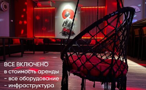 Арендовать Лофт ИЗНАНКА. Зал ТОКИО. Пространство с затемненным освещением, красные световые акценты на стенах. Мебель: черные скамьи вдоль стены, плетеное подвесное кресло с красной подушкой. На стене картина с изображением женщины. На полу паркет. Текст: "Все включено в стоимость аренды - все оборудование, инфраструктура".
