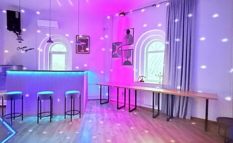 Арендовать Центрум. Уютное светлое пространство круглой формы. Просторная комната с барной стойкой слева, тремя высокими стульями, длинным деревянным столом справа. Арочные окна, занавески, светодиодная подсветка розового и синего цветов, паркетный пол, современные постеры на стенах.
