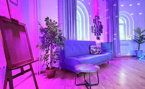 Арендовать Центрум. Уютное светлое пространство круглой формы. Просторная комната с деревянным полом, синим диваном и круглым столиком. На стене фотографии, светодиодная подсветка розового и голубого цветов. Растения в кадке и мольберт. Арочные окна с серыми шторами.