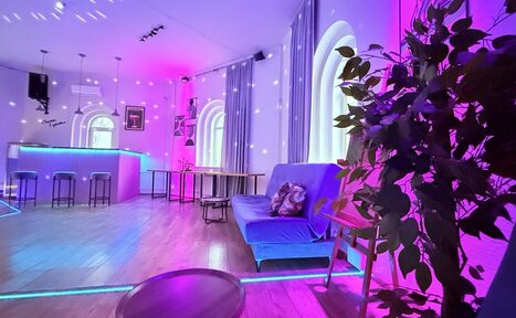 Арендовать Центрум. Уютное светлое пространство круглой формы. Просторная комната с барной стойкой, высокими табуретами и диваном. Окна арочные, пол деревянный. Освещение разноцветное, в основном пурпурное. На стенах и потолке световые эффекты. В углу растения, столы и стулья.