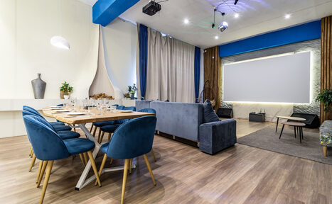 Арендовать Rubin Loft (Рубин Лофт). Зал Сицилия. Современное пространство с деревянным полом, обеденным столом и синими стульями. Серая мягкая мебель рядом с проектором и экраном. Потолочные светильники и декоративные элементы. Светлые шторы, зелёные растения на фоне.