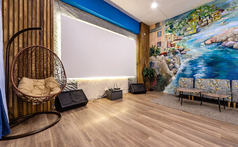 Арендовать Rubin Loft (Рубин Лофт). Зал Сицилия. Просторная комната с деревянным полом. На стене роспись с пейзажем. Имеется проектор, пара колонок, висящее кресло-кокон, пара стульев с серой обивкой и растительность. Стена оформлена под дерево, освещение точечное и диодное.