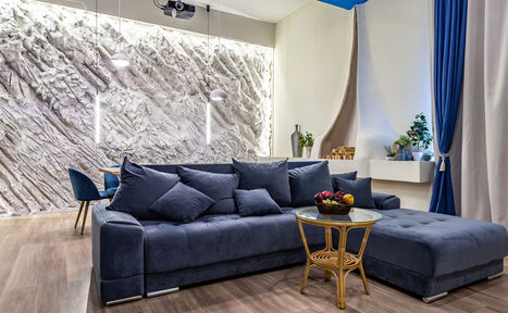 Арендовать Rubin Loft (Рубин Лофт). Зал Сицилия. Современное пространство с серым угловым диваном, круглым стеклянным столиком на деревянных ножках, декоративной стеной с подсветкой, подвесными светильниками, синими шторами, деревянным полом и белыми стенами.