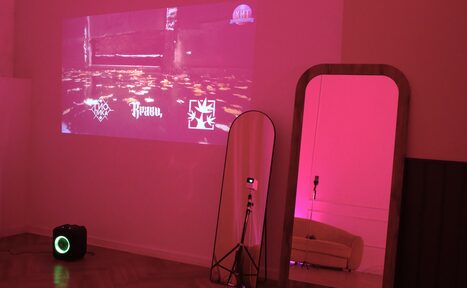 Арендовать Стильный лофт для уютных вечеринок. Комната с проектором на стене, показывающим красочное изображение. На полу черная колонка. Два больших зеркала, одно с деревянной рамой, другое на треноге. Слабое розовое освещение, диван виден в отражении зеркала. Паркетный пол.