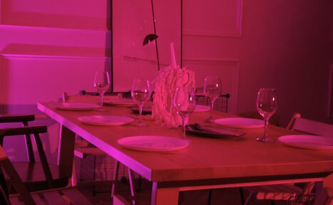 Арендовать Стильный лофт для уютных вечеринок. Столовая в розовом освещении, деревянный стол с шестью накрытыми приборами, бокалы для вина, декоративная композиция по центру. Стулья разных дизайнов вокруг стола. Белые наклонные панели на стенах.