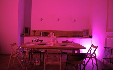 Арендовать Стильный лофт для уютных вечеринок. Столовая зона с деревянным столом, окруженным разными стульями, на столе тарелки и бокалы. На заднем плане открытые полки с предметами и декоративное освещение розового цвета. Белые стены, паркетный пол. Трековые светильники на стене.