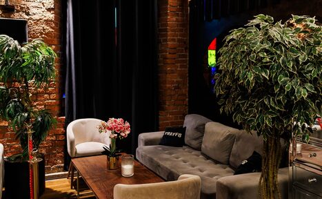 Арендовать Двухуровневая event-площадка 550 м². Лофт-пространство с кирпичными стенами, серым диваном, креслом, деревянным столом, растениями в горшках. Тёмные шторы, мягкое освещение, узорчатый ковёр. На столе свеча и цветы. Подушка с надписью «TREFF».