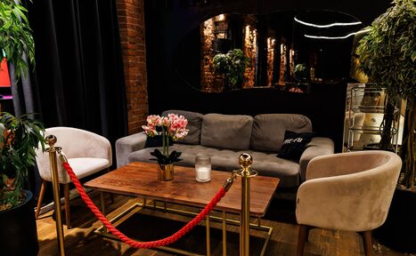 Арендовать Двухуровневая event-площадка 550 м². Лаунж пространство с диваном и двумя креслами вокруг деревянного стола на золотых ножках. Красный канат на стойках создает барьер. На столе свеча и цветы в горшке. На стене темное зеркало. Кирпичная стена и деревянный пол. Тёмные шторы слева.