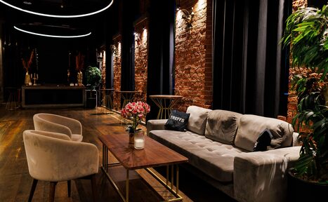 Арендовать Двухуровневая event-площадка 550 м². Интерьер просторного помещения с кирпичными стенами и деревянным полом. Зона отдыха с серым диваном, двумя бежевыми креслами, деревянным столом и декором. Потолочные светильники в виде кругов, текстиль тёмных тонов, зелёные растения.