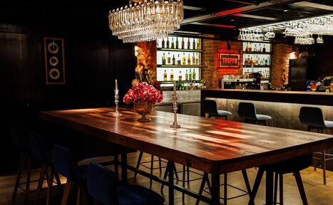 Арендовать Двухуровневая event-площадка 550 м². Барное пространство с деревяным столом и синими стульями. Над столом висят кристаллические светильники. На столе декор из цветов и свечей. На заднем плане барная стойка с высоким стульями и полки с бутылками. Стены из красного кирпича.